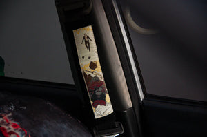 In a close-up of a car's interior, a seatbelt displays an artistic design inspired by the Jujutsu Kaisen Mystery Pack from Incompletegl. The abstract elements, featuring a human figure and various shapes, create intrigue against the partially blurred car upholstery and window.