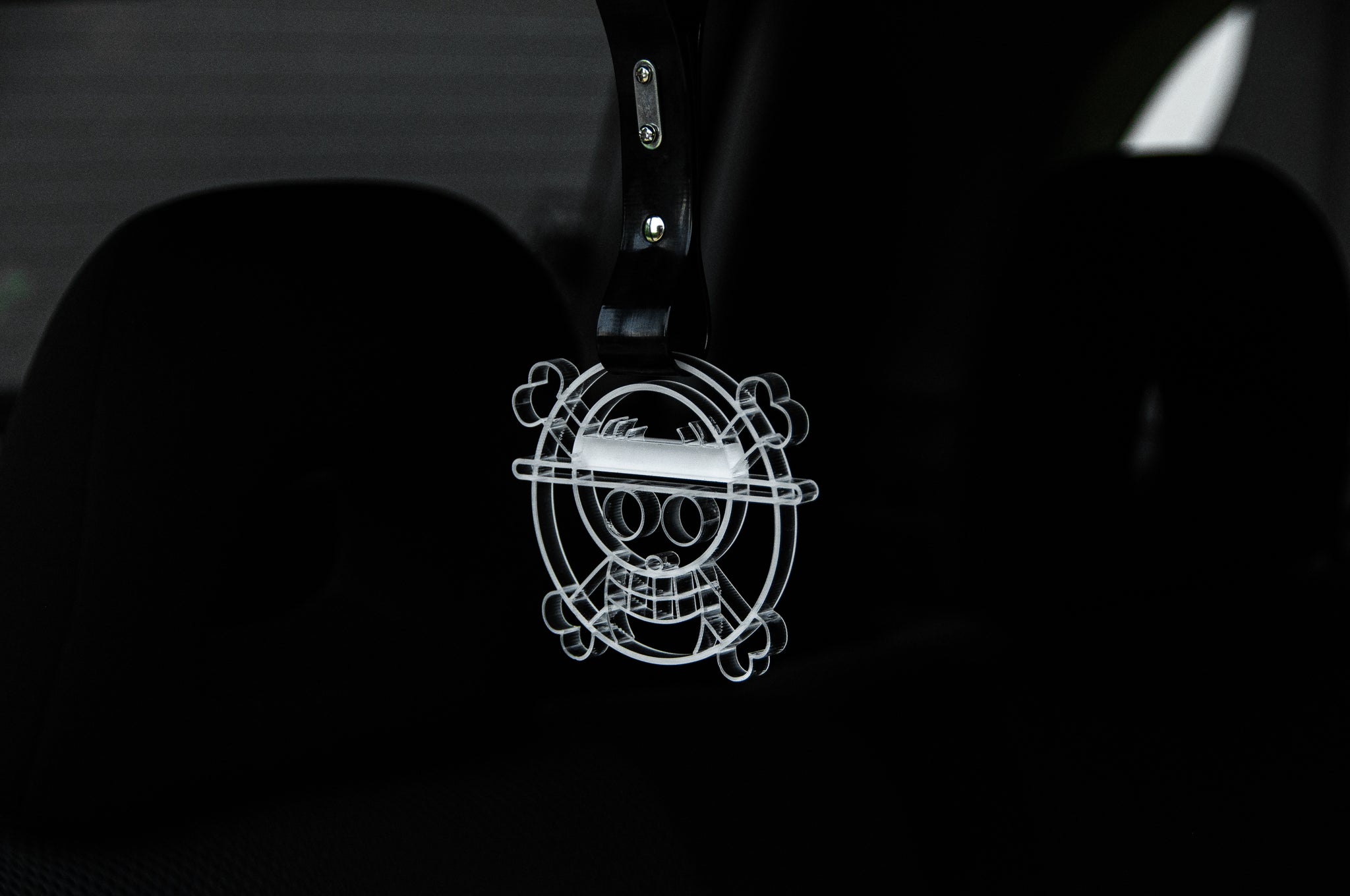 The Straw Hat! (Jolly Roger) | Tsurikawa! by Incompletegl is a transparent pendant featuring a cartoon skull with a straw hat and crossbones, elegantly dangling from a tsurikawa strap in the car interior, with the dark background hinting at the car seat for an edgy touch.