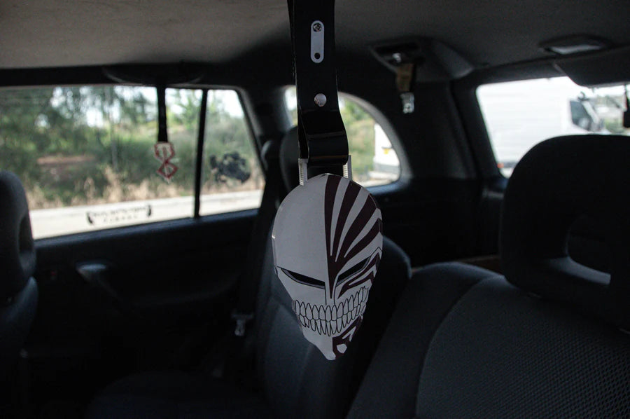 An anime-themed mask from the Bleach Mega Mystery! | Anime Bundle Pack by Incompletegl, featuring black and white stripes, hangs in the backseat of a car, suspended from the ceiling. The interior is grey, while the view through the window reveals the unfolding outside world.