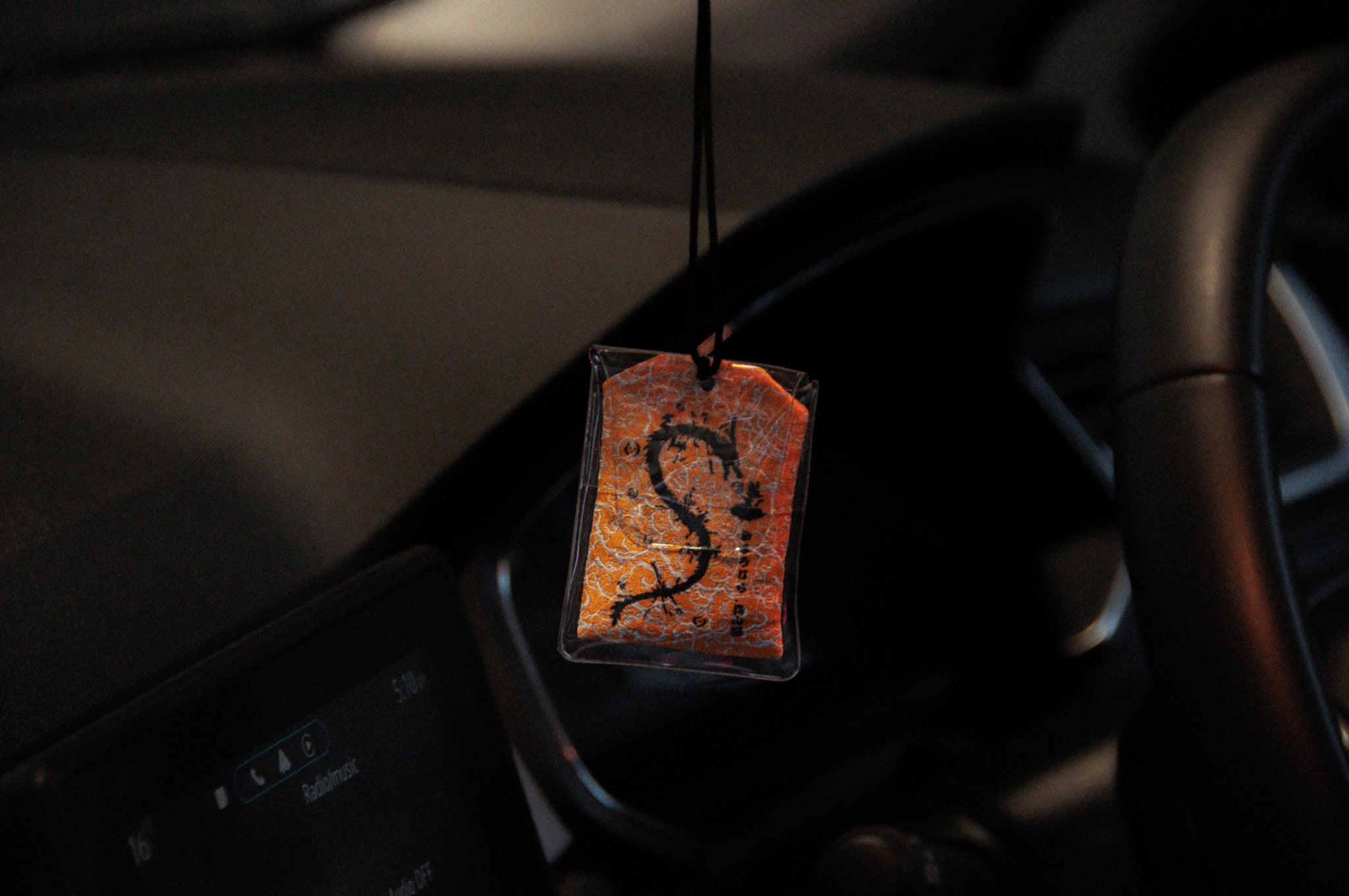 A close-up of a car's interior showing an Incompletegl Goku x Shenron Omamori Charm hanging from the rearview mirror. The charm features a letter "S" printed on a patterned orange background. The dashboard and steering wheel are partially visible in the background.