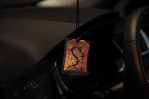 The Goku x Shenron | Omamori Charm by Incompletegl is a hanging rectangular ornament featuring intricate orange patterns and a decorative "S" in the center. It is suspended from the rearview mirror of a car, with parts of the dashboard, steering wheel, and infotainment system visible in the background.