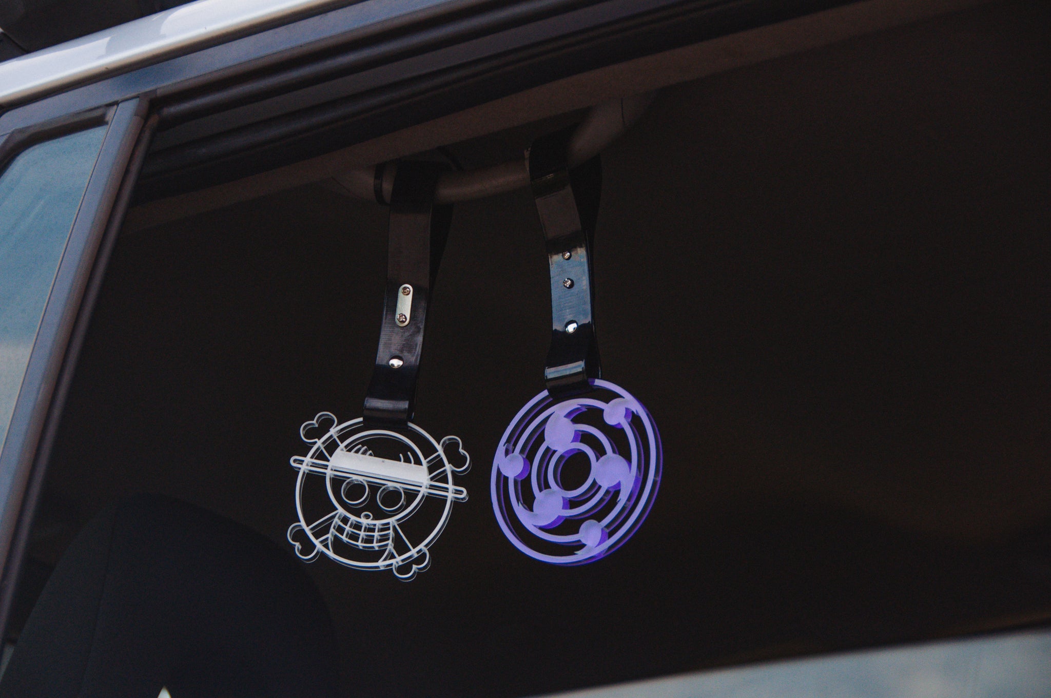 Two car window hangings: one with a stylized skull wearing a hat, and the other showcasing a Rinnegan Tsurikawa pattern. These Incompletegl "Rinnegan! | Tsurikawa" accessories are mounted on black straps inside a partially open car window, infusing JDM style.