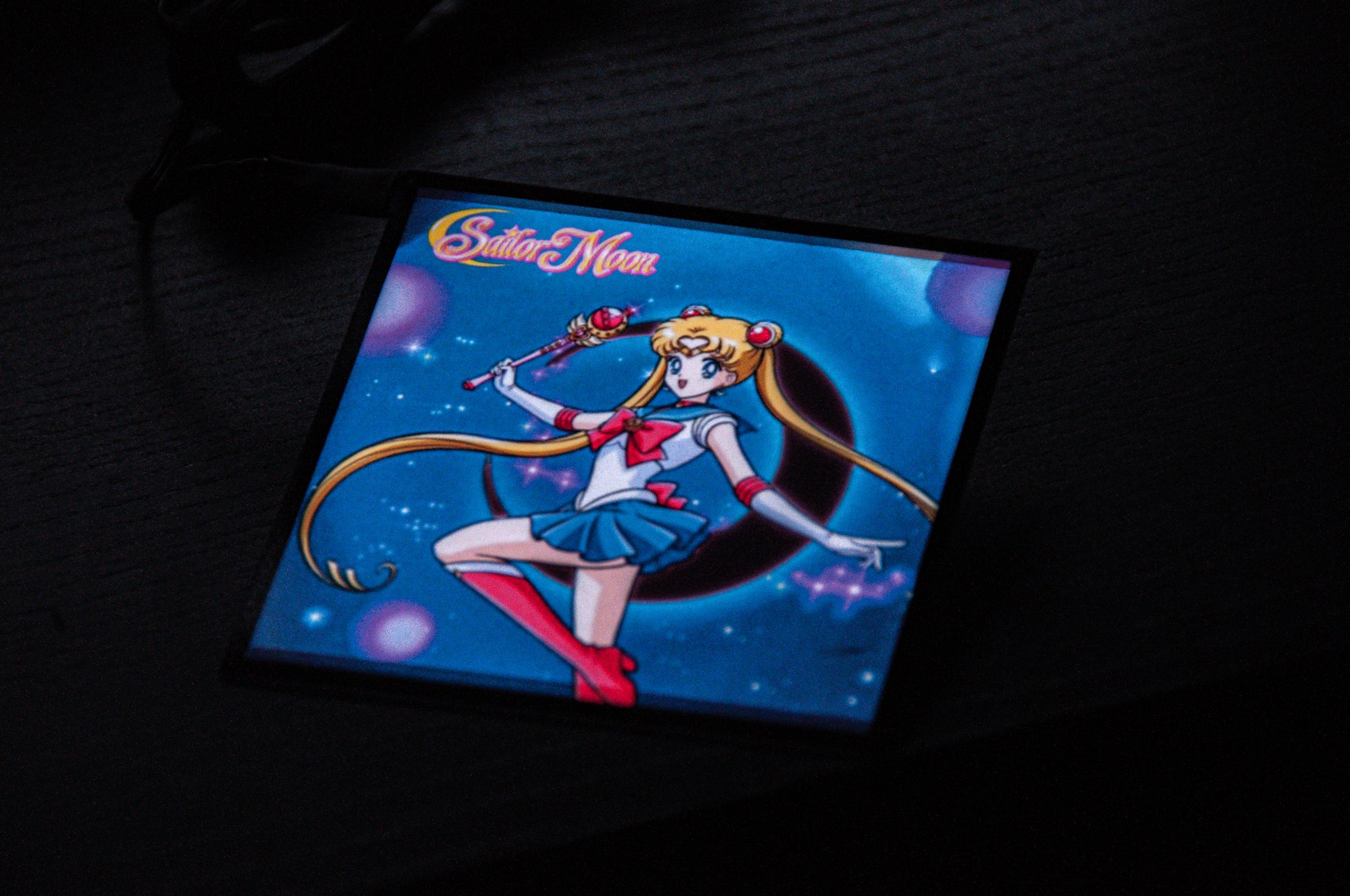 An image of the anime character Sailor Moon is vibrantly displayed on the square screen of the black Sailor Moon! | Glow Panel by Incompletegl. Sailor Moon wields a wand in her right hand, with her left arm extended, and she stands in her iconic sailor suit against a dark, starry backdrop illuminated by glowing orbs—making it an ideal JDM accessory for any car interior.