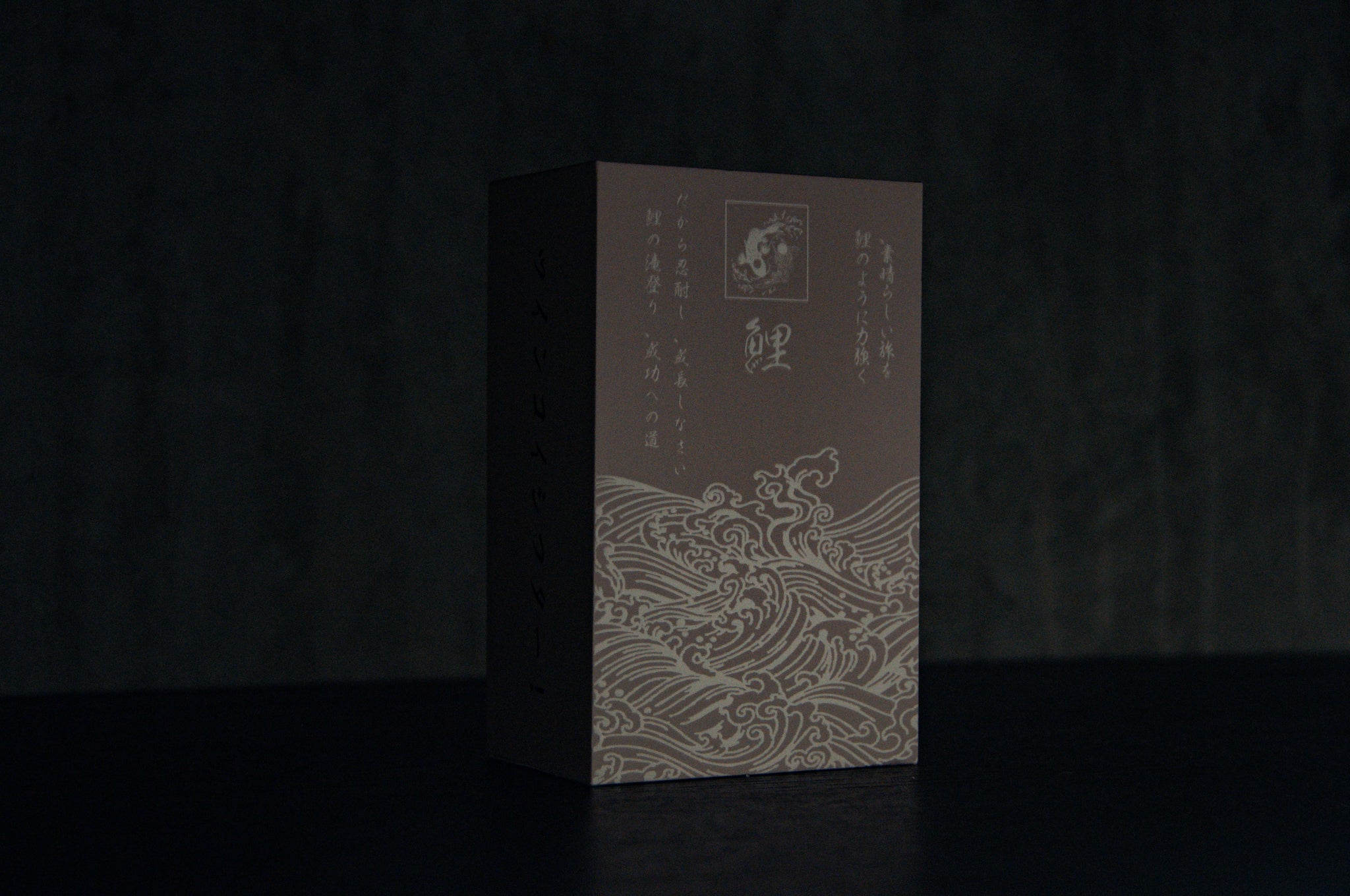 A dark, upright rectangular box against a dim background showcases the Twin Koi Fish | Artisan Shift Knob from Incompletegl. It features elegant wave line art and Japanese text, capturing handmade artistry and reflecting simplicity and sophistication rooted in Japanese heritage.