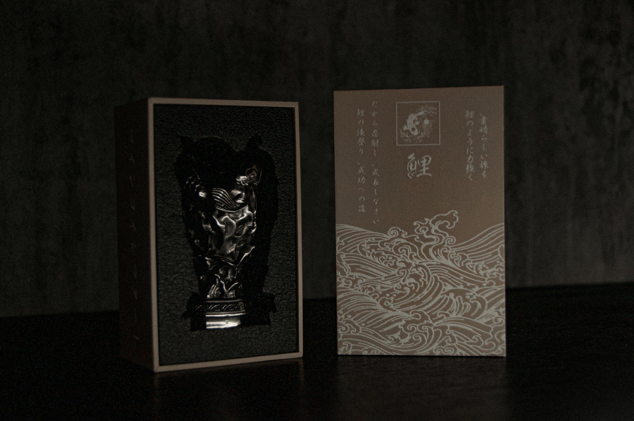 The Twin Koi Fish Artisan Shift Knob by Incompletegl, resembling an animal face with Japanese flair, is partially revealed inside a dark box featuring intricate Japanese text and designs. Beside it is packaging adorned with calligraphy and a wave pattern, showcasing exquisite handmade craftsmanship.