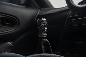 Close-up of a car gear shift showcasing the Twin Koi Fish | Artisan Shift Knob by Incompletegl. The dimly lit interior reveals part of the steering wheel and dashboard, emphasizing its unique handcrafted JDM style.