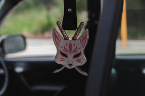 Inside the car, an interior scene features a hanging air freshener from **Incompletegl** called **Kitsune Mask! | Tsurikawa!**, designed with pink markings that evoke Japanese folklore. Behind it, two miniature swords are crossed, and the blurred scenery of the road and greenery is visible in the background.