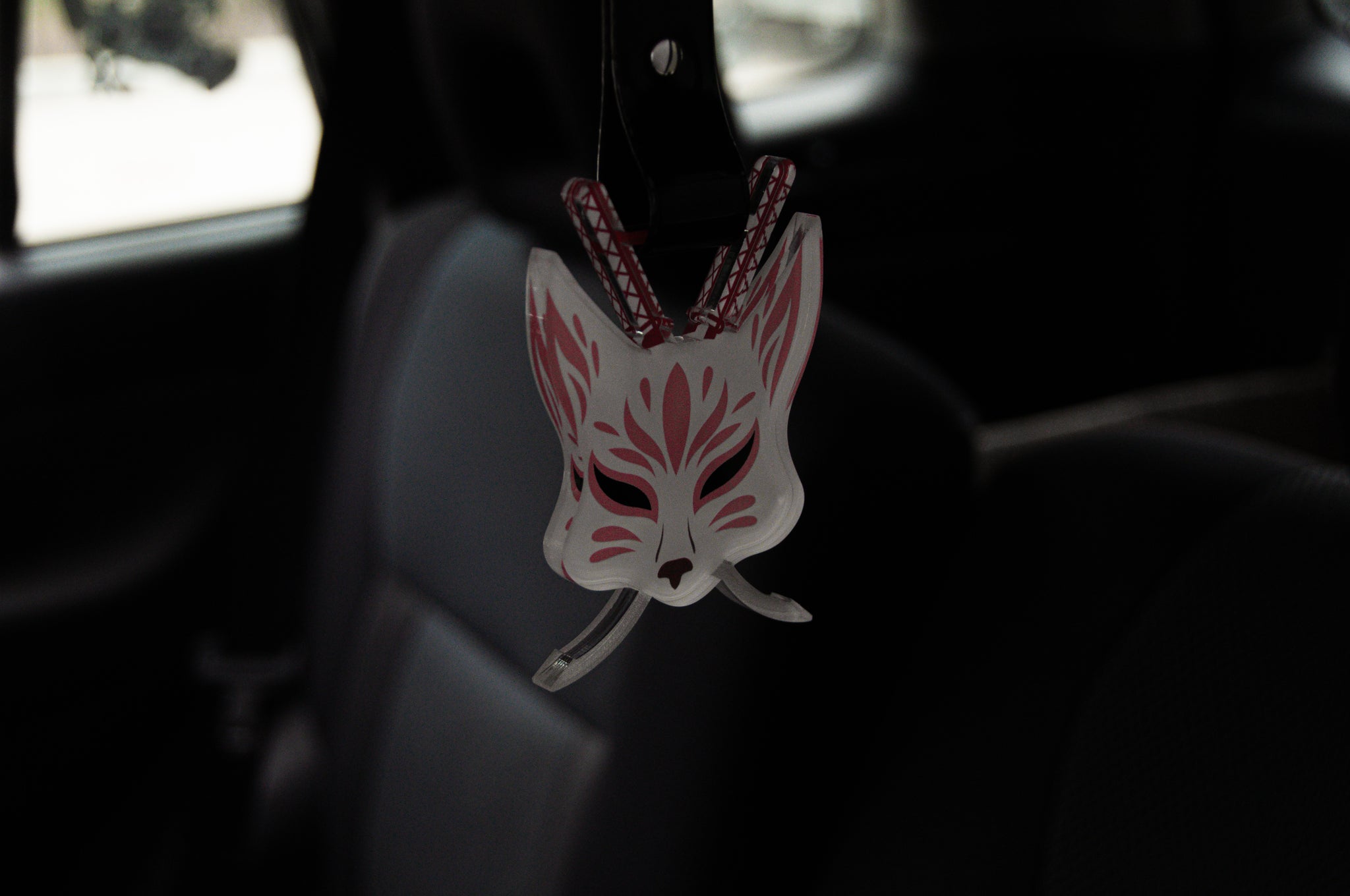 An Incompletegl Kitsune Mask in white, adorned with pink markings, hangs from an Incompletegl Tsurikawa, infusing the dimly lit car interior with an aura of Japanese folklore.