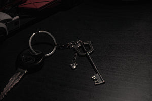 A metal key with a circular keychain, adorned with an Anime Keychain charm from the Anime Keychain Pack! | Bundle Pack by Incompletegl, lies on a dark surface. The keys are partially illuminated, casting subtle reflections, while the background remains dimly lit.