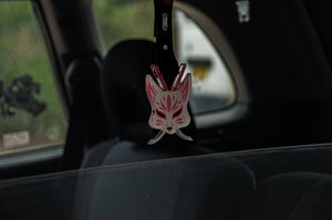 A pink and white Kitsune Mask! | Tsurikawa! from Incompletegl elegantly hangs from a car's rearview mirror, evoking the essence of Japanese folklore. The dark car interior contrasts with the blurry green and white scene outside, imparting a mystical charm to every journey.