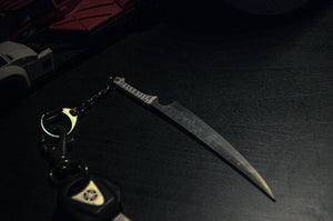 A close-up photo showcases the Incompletegl Zangetsu Keychain, which features a decorative mini-dagger that evokes the design from Bleach. Also attached is a set of car keys, including a black and silver key fob emblazoned with a triangular logo. The items are placed on a dark surface with a red object partially visible in the background.