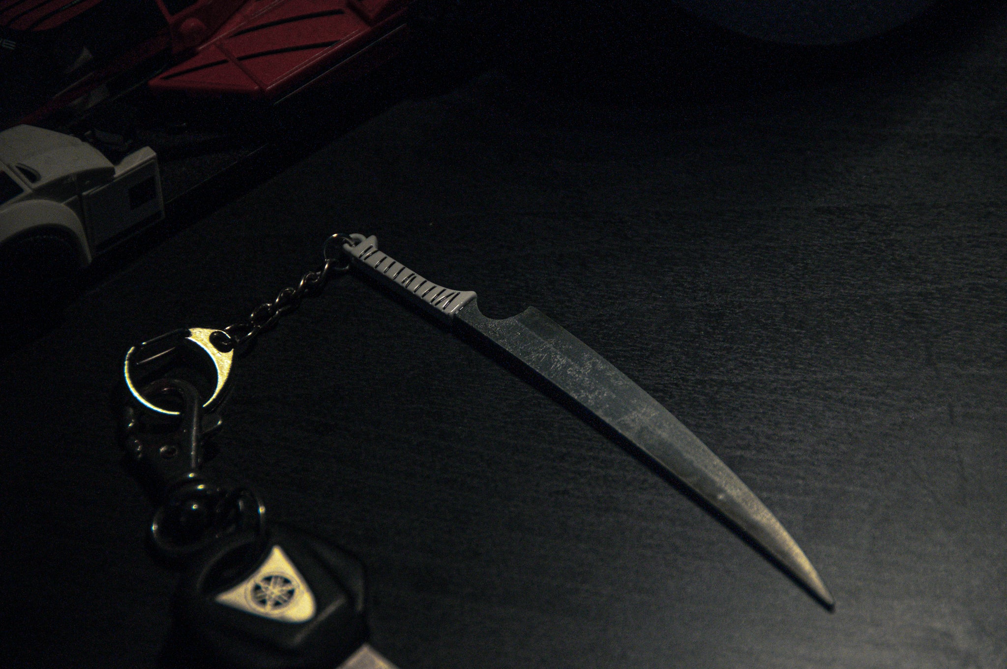 A close-up shot features a dark metal knife with a serrated handle from the Anime Keychain Pack! | Bundle Pack by Incompletegl, resting on a dark surface. The keychain also holds a car key and a metallic keyring. Nearby, there's a partially visible red and white object, possibly toy vehicles.