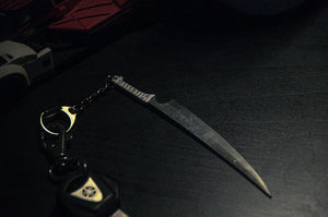 In a dimly lit, atmospheric image, an anime-inspired knife-shaped keychain rests on a dark surface. To the left, a keyring holds car keys and a handcuff charm, subtly suggesting it might be part of the Bleach Mega Mystery! Anime Bundle Pack from Incompletegl. In the background, you can faintly see a toy car.