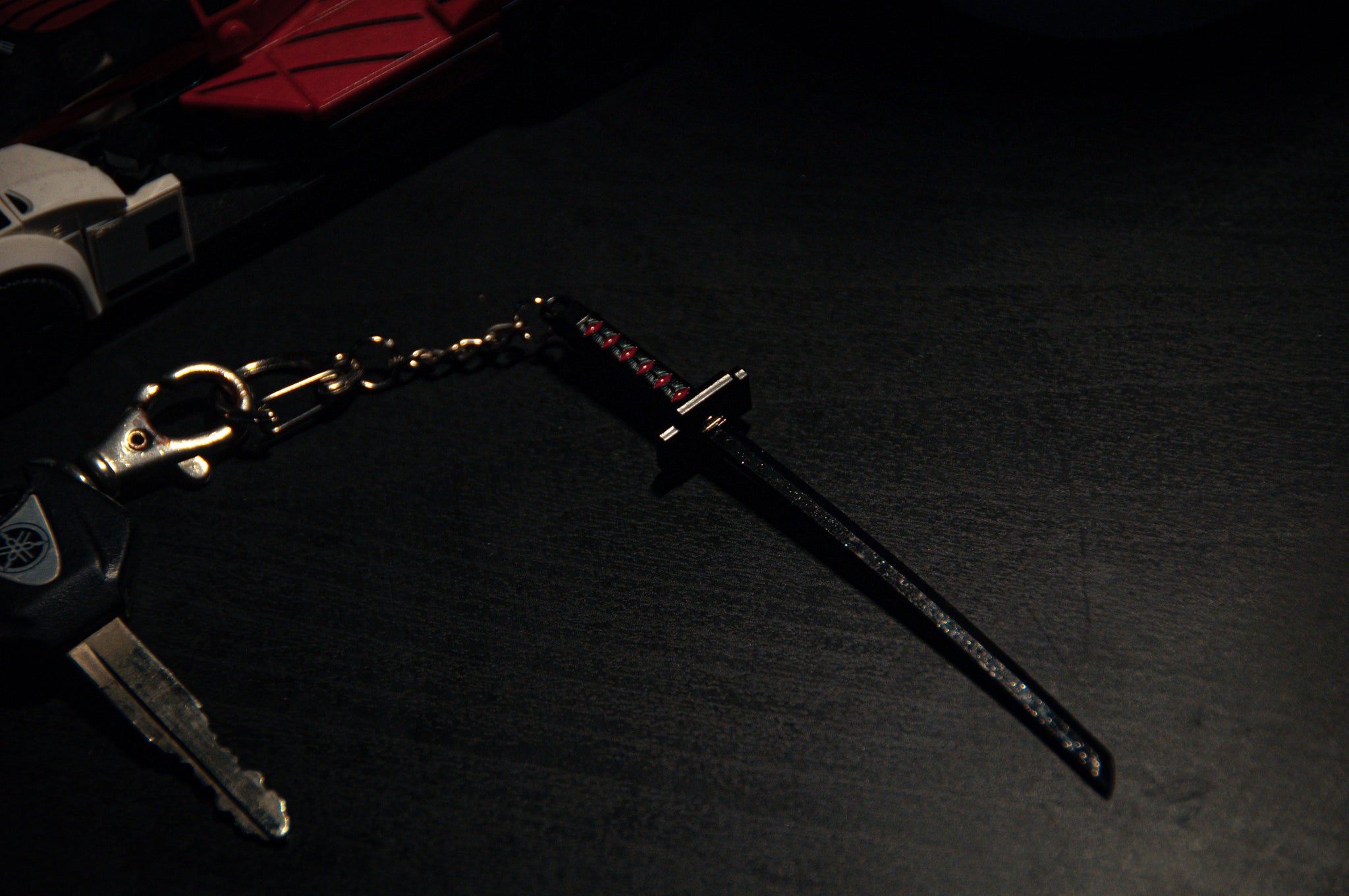 A close-up of a set of keys on a dark surface. The Anime Keychain Pack! | Bundle Pack by Incompletegl includes a miniature katana sword reminiscent of Bleach Zangetsu, featuring a black scabbard and a decorative handle. Beside the keys, there is a red toy car partially visible.