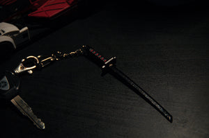 A car key with a Yamaha logo and an Incompletegl Tensa Zangetsu Keychain sit on a dark surface. The keychain, inspired by the sword from Bleach, features red details on the hilt and is connected to the key by a gold chain and clip. In the background, partially visible, are toy cars.