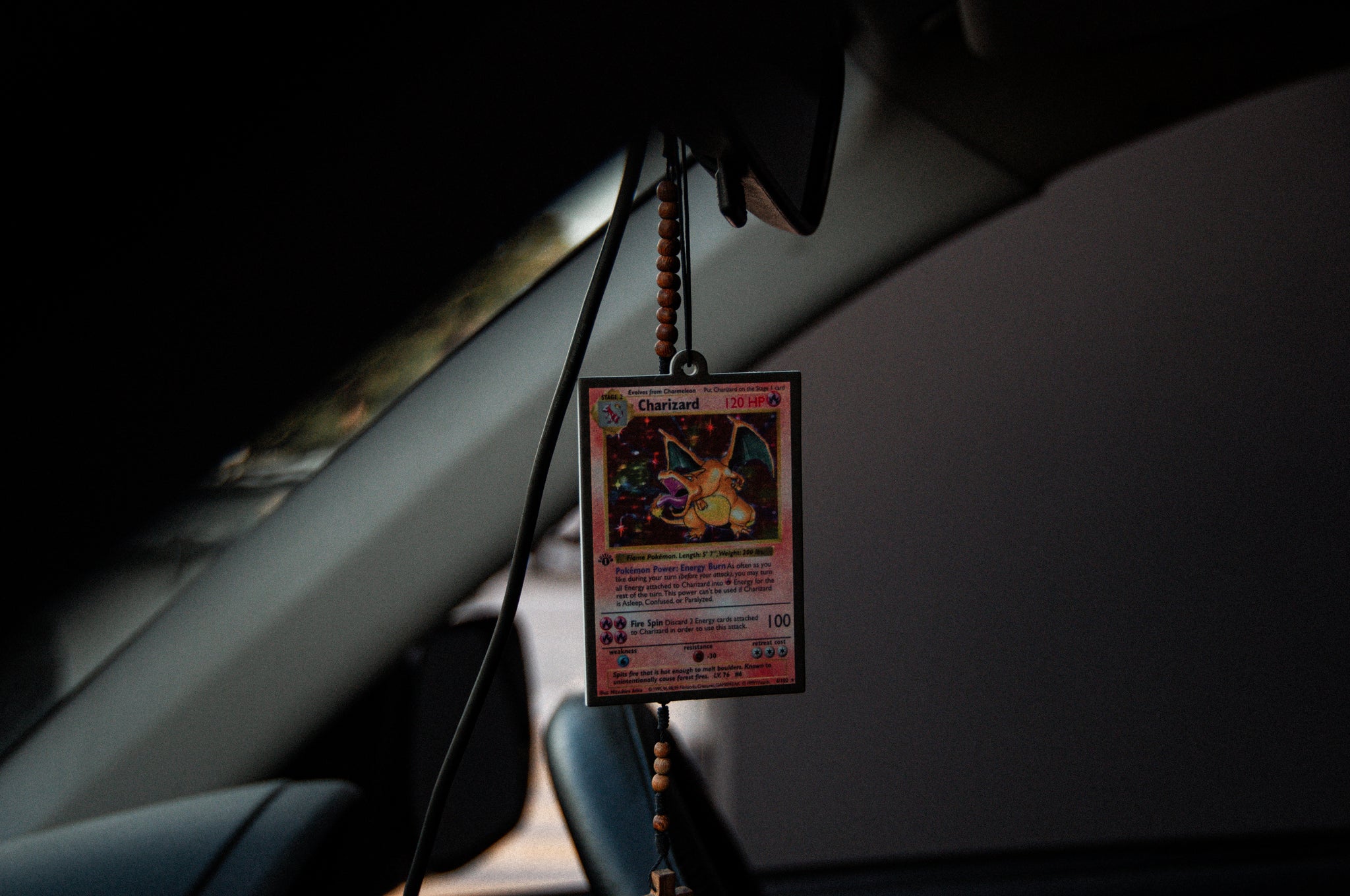 A JDM-inspired Charizard! Air Freshener by Incompletegl hangs from a beaded necklace on the rearview mirror, serving as both decoration and air freshener. The sharply focused product stands out against the dimly lit car interior, adding a stylish touch to the ride.