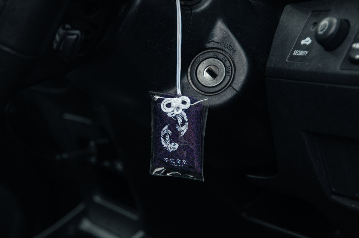 A close-up of a car's ignition area features a key in the ignition slot. Hanging from the key is a small, purple fabric charm with white embroidery depicting two koi fish in a circular pattern. The charm’s white string has an intricate knot, complementing the cozy feel provided by the Koi Fish Comfort Pack from Incompletegl nearby.