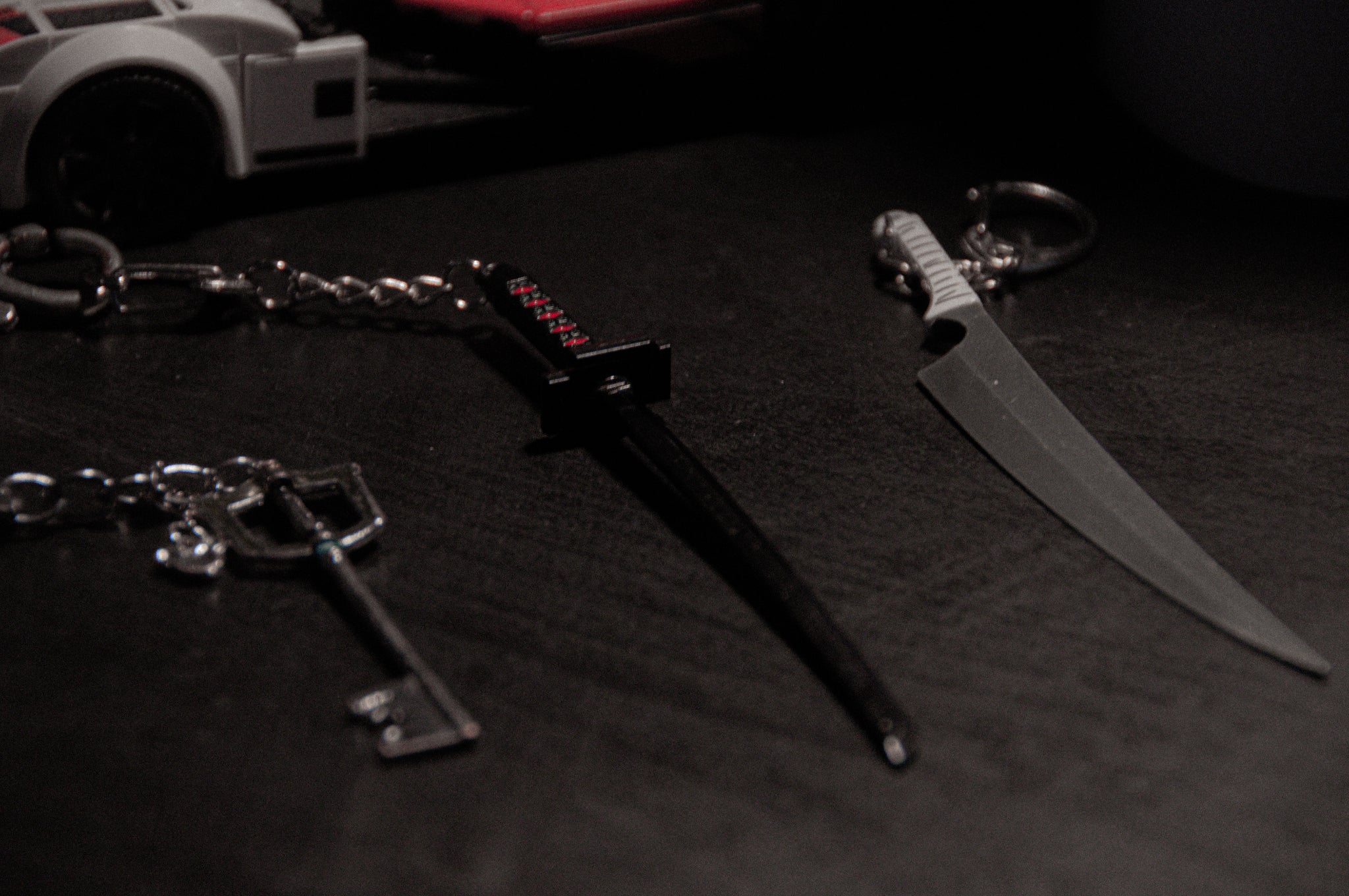 Close-up of three keychains from the "Anime Keychain Pack! | Bundle Pack" by Incompletegl on a dark surface. The left keychain resembles the Kingdom Hearts Keyblade, the middle one showcases Bleach's Zangetsu sword, and the right one is shaped like a large knife. A partial view of a white toy car with red accents can be seen in the background.