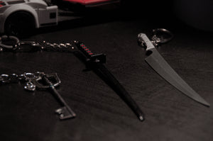 Close-up image of three metallic keychains from the Anime Keychain Pack! | Bundle Pack by Incompletegl, arranged on a dark surface. They are shaped like different types of weapons: a Kingdom Hearts Keyblade, a katana sword reminiscent of Bleach Zangetsu, and a curved knife. A partial view of a white model car with a red section is visible in the background.