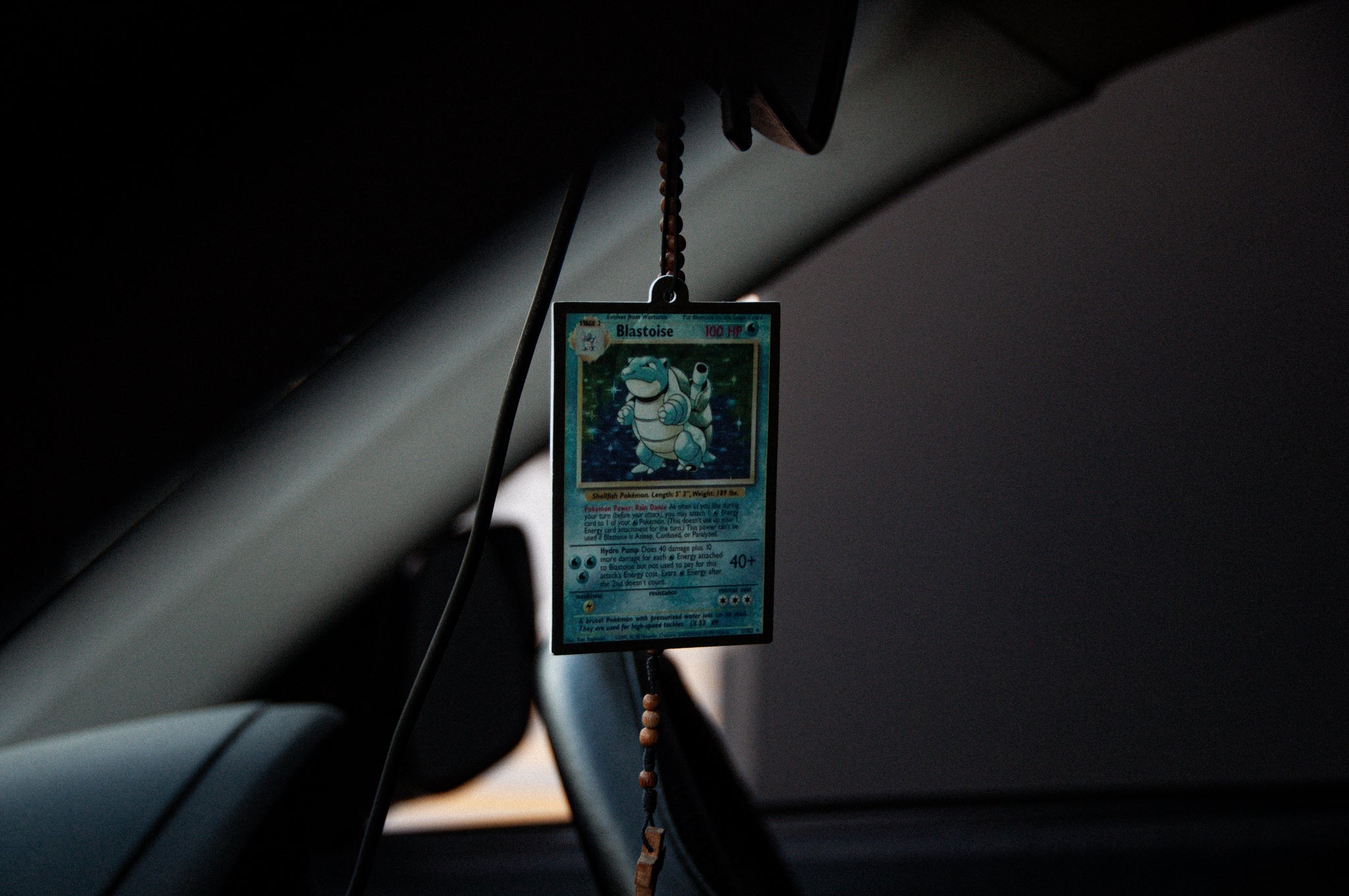 An Incompletegl Blastoise!! air freshener, which doubles as a trading card, hangs from a string of beads near the car's rearview mirror. Encased in protective plastic, it discreetly eliminates odors while serving as the centerpiece in the dimly lit interior.