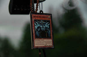 A close-up of the "Obelisk the Tormentor" air freshener by Incompletegl, showcasing its intricate design from the "Yu-Gi-Oh!" series. The air freshener is suspended from a beaded string, likely within a car, and encased in a protective plastic cover. The blurred background outside the car reveals green foliage.