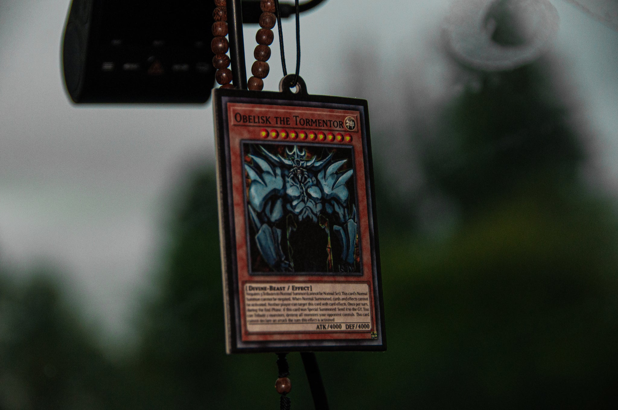 An "Obelisk the Tormentor" Yu-Gi-Oh! card from the Ultimate Deck! | Air Freshener Bundle by Incompletegl dangles on a beaded chain beside a watermelon-scented air freshener, serving as a car ornament. The blurred background reveals lush green foliage outside the car.