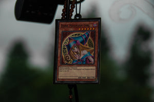 Close-up of a "Dark Magician Girl" trading card from the Ultimate Deck! | Air Freshener Bundle by Incompletegl, encased in a plastic holder and hanging from a string of beads. The background is blurred to focus attention on the card, which is featured alongside an Obelisk the Tormentor figurine.