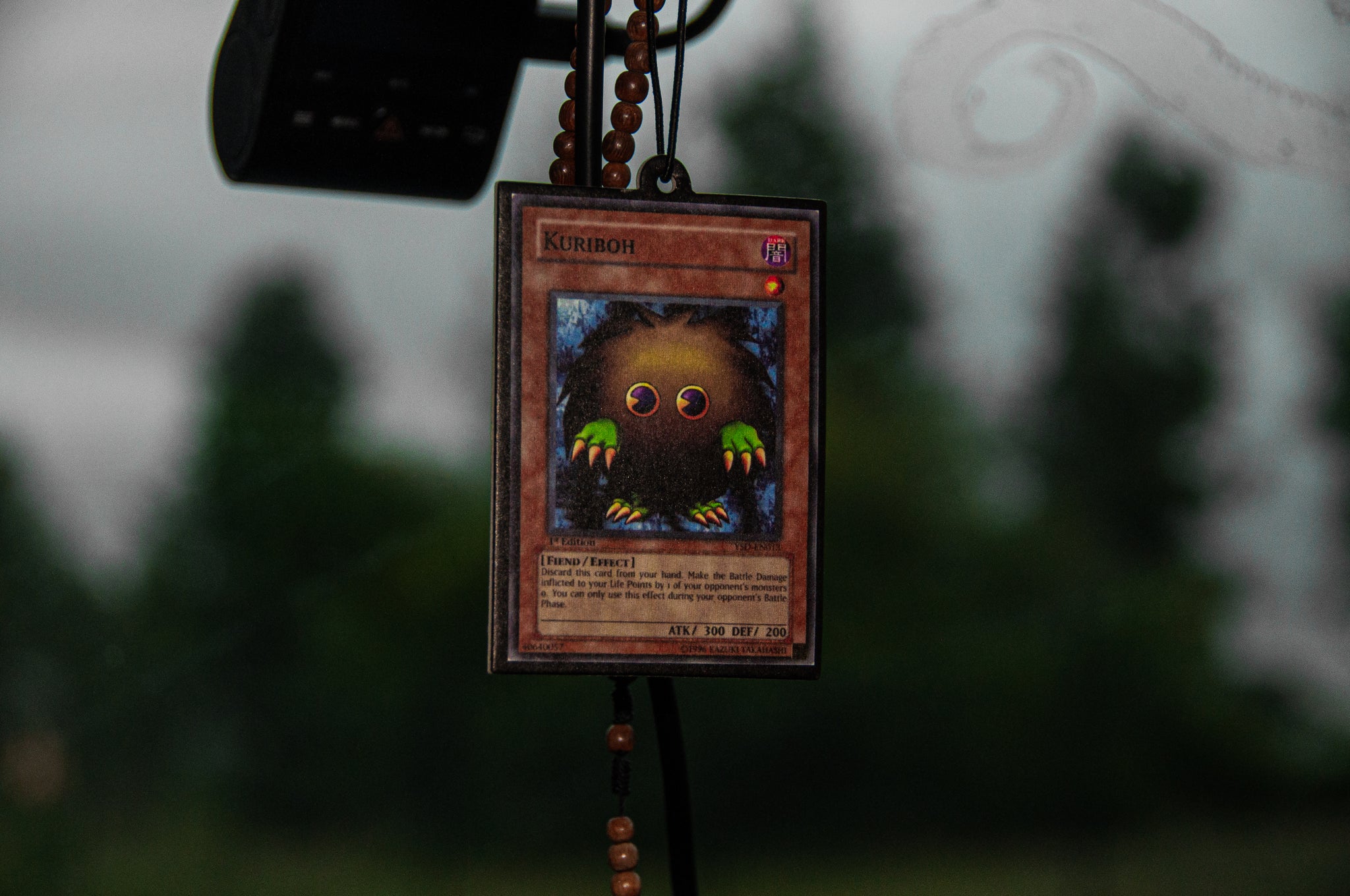 Hanging from the rearview mirror is a Kuriboh! air freshener by Incompletegl, adorned with a beaded necklace. The air freshener depicts a small, fuzzy, brown creature with big eyes and colorful claws, known for its defensive abilities in the Yu-Gi-Oh! series. The background showcases blurred greenery.