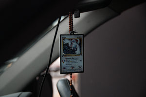 An air freshener from Incompletegl featuring the Lugia!! design hangs gracefully from a beaded chain on the rearview mirror, paired with a black ice scent. The card is encased in a protective sleeve, swaying subtly as the car moves, with its background elegantly blurred.