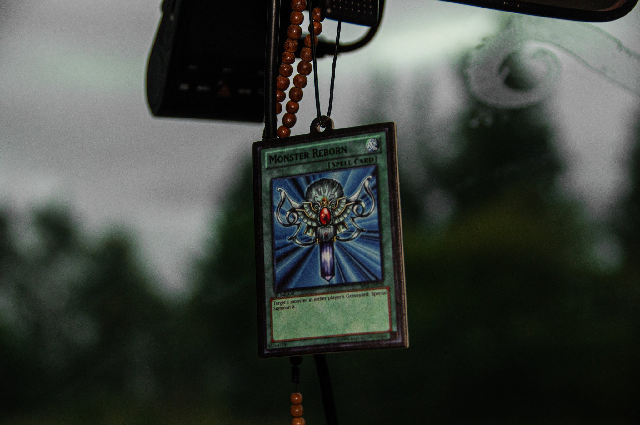 A Monster Reborn! air freshener by Incompletegl hangs from a rearview mirror using a beaded chain. The background shows a blurred view of trees and a cloudy sky, while the freshener, prominently featured in the foreground, is encased in a protective sleeve.