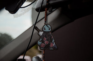 A car interior showcasing an anime car accessory from Incompletegl, hanging from the rearview mirror. The Squirtle x Akatsuki Air Freshener charm features a cartoon turtle dressed in a ninja outfit with red flower patterns. In the background, the road appears as a blur.