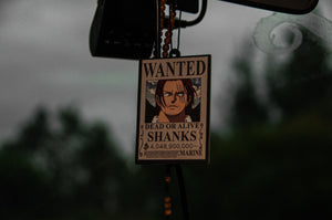 A Shanks Wanted Poster! Air Freshener from Incompletegl, featuring an animated image of Shanks from the Red-Haired Pirates with a bounty of 4,048,900,000 berries, hangs from a car rearview mirror. The background is blurred, showing green foliage outside the window.