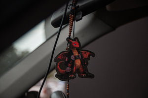 An air freshener named "Charizard x Akatsuki!," by Incompletegl, featuring a black ice scent and inspired by the Charizard design, hangs from a rearview mirror. The red and black hues stand out in the dim interior, accented with a beaded necklace that enhances its mystical appeal.