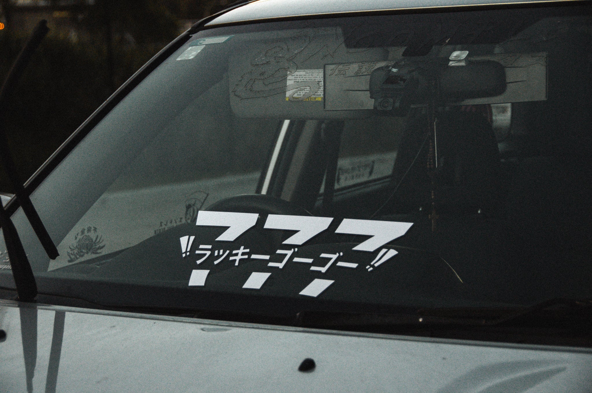 A car windshield adorned with a "Lucky 777!! | Mini Banner" from Incompletegl, accompanied by Japanese text beneath it. The dashboard features a camera mounted near the rearview mirror, and various small decals on the glass add character. The setting appears to be outdoors in low lighting.