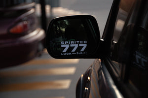 A car side mirror featuring the "Spirited 777! | Mini Decal" from Incompletegl catches the eye, reminiscent of a Japanese charm. In the background, another car's rear and a yellow-striped road surface are visible. The overall scene has a dimly lit ambiance.