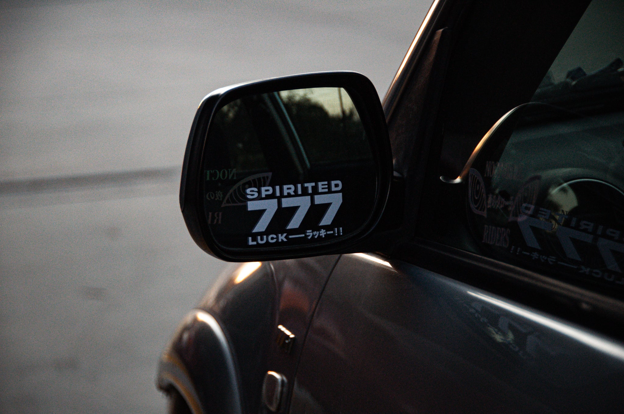 Close-up of a car's side mirror displaying a "Spirited 777! | Mini Decal" by Incompletegl that reads "SPIRITED 777 LUCK ラッキー!!" The reflection shows part of another sticker on the car window. The background is a blurred, lightly lit outdoor setting, evoking the feel of a Japanese charm.