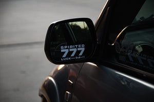 Close-up of a car's side mirror displaying a "Spirited 777! | Mini Decal" by Incompletegl that reads "SPIRITED 777 LUCK ラッキー!!" The reflection shows part of another sticker on the car window. The background is a blurred, lightly lit outdoor setting, evoking the feel of a Japanese charm.