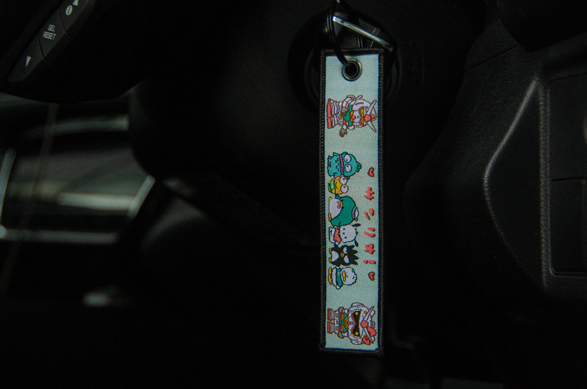 A Jet Tag from the Incompletegl collection, featuring Sanrio x Gundam characters, hangs from a vehicle's ignition switch. The colorful, vertically aligned figures are attached to a black key. The car's interior, including the dashboard and steering wheel, is visible, showcasing playful mecha aesthetics that complement the JDM Jet Tag vibes.
