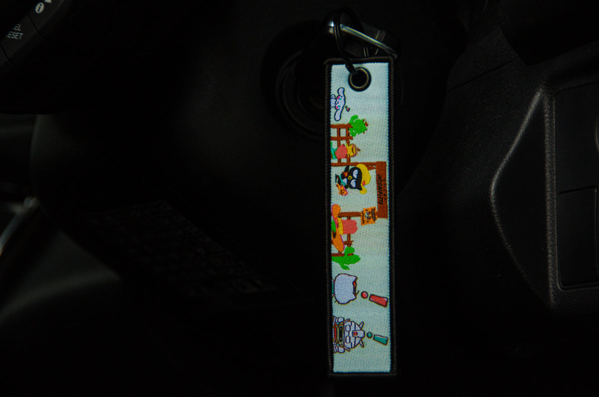 A close-up photo of a Sanrio x Gundam! Jet Tag keychain hanging from an ignition in a car. The keychain, by Incompletegl, is rectangular and features colorful illustrations of various characters and objects including a penguin, a sheep, and a robot with mecha aesthetics set against a light background.