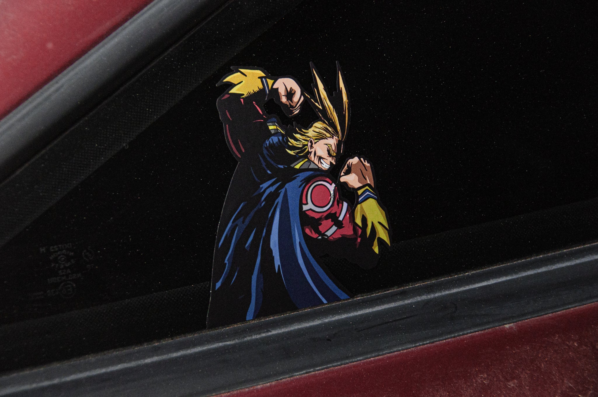 A close-up of a car window featuring the ALL MIGHT! | Holographic Peeker Decal from Incompletegl, depicting All Might from My Hero Academia with blonde hair, dressed in a red and blue superhero costume, fists raised in a fighting pose against a black background. The car’s exterior is red.