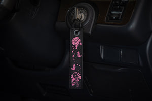 An elegant pendant from the Geisha Mega Pack! by Incompletegl, featuring intricate pink cherry blossom tree designs, petals, and a traditional Japanese umbrella on a black base, hangs gracefully from the car's ignition switch. Ideal for car enthusiasts with a taste for refined JDM accessories, it adds a touch of sophistication with the dashboard visible in the background.