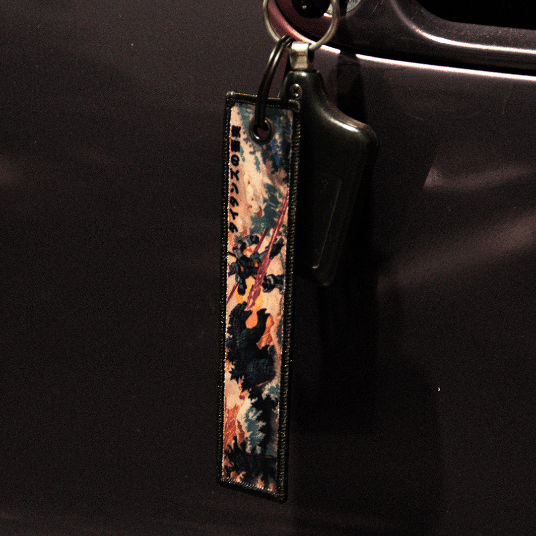 A close-up of a car key inserted into a car door lock reveals a rectangular fabric keychain with a vibrant design, showcasing an aircraft and cloud patterns. This keychain is inspired by human technological advancements and features the text "remove before flight." It is the Godzilla x Gundam Jet Tag by Incompletegl.