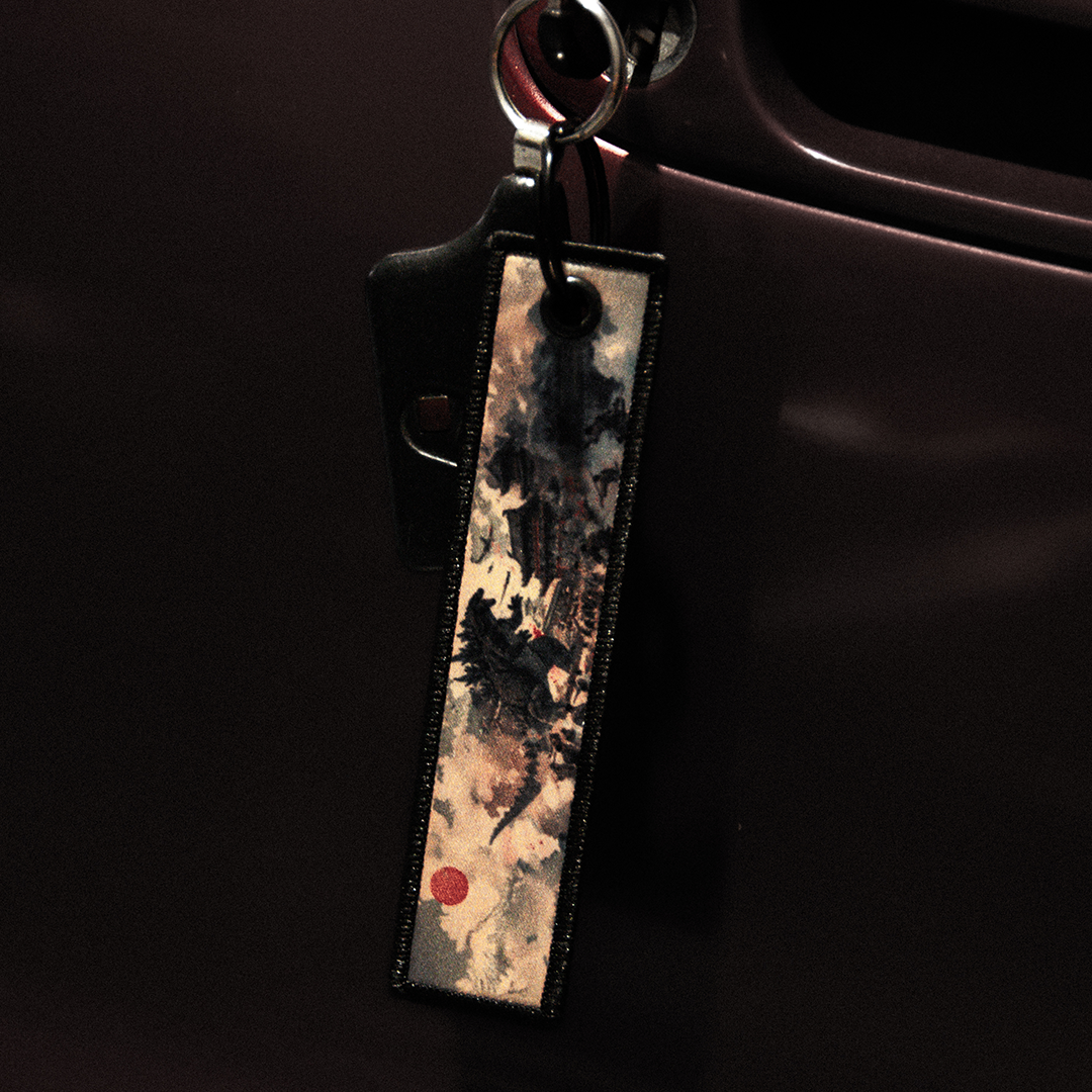 A close-up of a car key inserted into a lock. The keychain proudly displays the Godzilla x Gundam Jet Tag by Incompletegl, adorned with an artistic design featuring watercolor-like brushstrokes and a small red circle in the bottom left corner evocative of Godzilla's fiery battles. The clean, dark, glossy car door reflects human technological advancements.