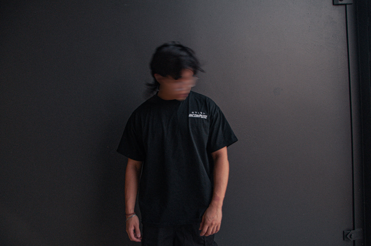 A person with blurred facial features stands against a dark gray wall, dressed entirely in black, featuring an all-black DONALD DUCK x RONIN SUIT Oversized T-Shirt from Incompletegl with text on the left side of the chest and matching shorts, exuding a contemporary ronin vibe. They are looking down with their head slightly tilted to the side.