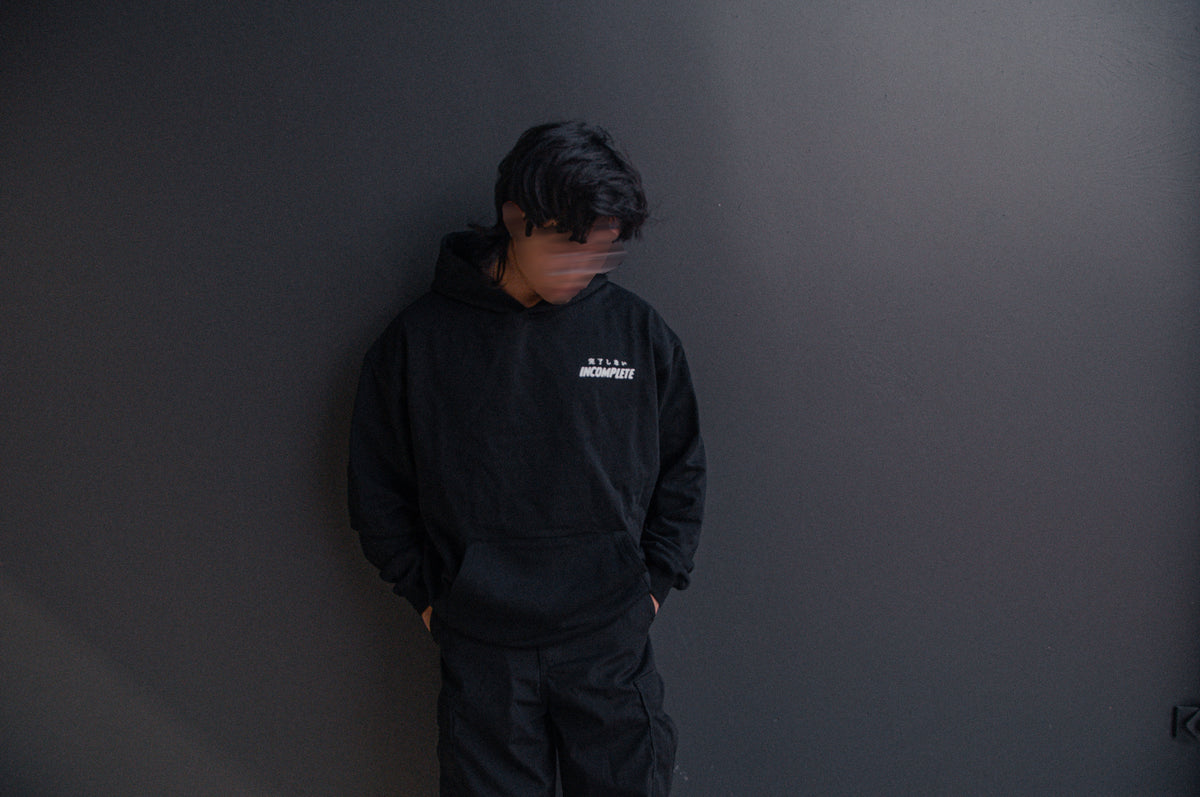 A person with dark hair, wearing an oversized GUNDAM x RONIN SUIT hoodie from Incompletegl and pants resembling modern samurai attire, stands against a dark gray wall. The individual's face is intentionally blurred, and their hands are in their pockets while looking downwards.