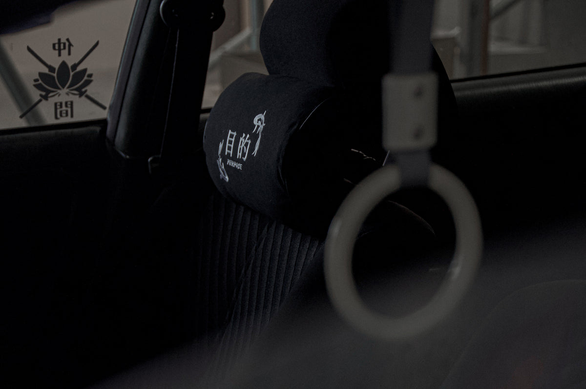 A close-up of a car's interior showcases the Koi Fish Pillows! headrest cushion from Incompletegl, featuring Japanese characters and a delicate design. In the foreground, a commuter handle dangles from the car's ceiling. The background is dimly lit, with part of a window visible, capturing the essence of Japanese culture.