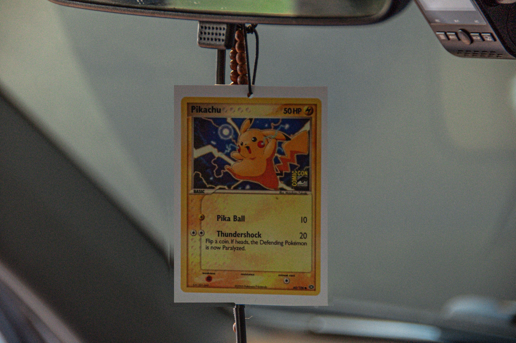 An Incompletegl Pokémon, Pikachu! air freshener is hanging from a rearview mirror inside a car. The air freshener features “Pikachu” at the top and lists its abilities "Pika Ball" and "Thundershock" with corresponding values. The background showcases part of the car's interior, highlighting the charm of having this Pikachu-themed decor.