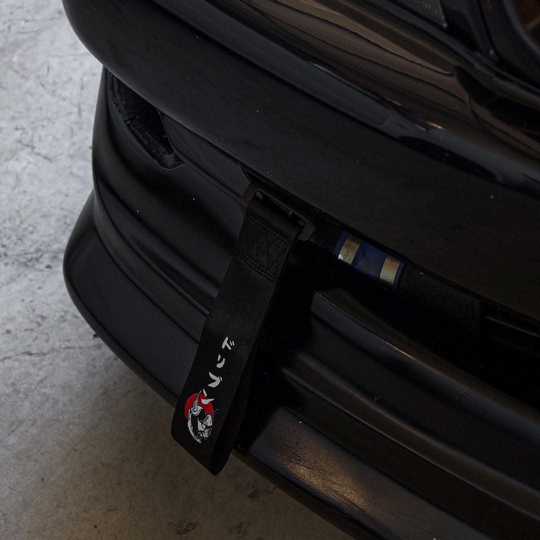 Close-up view of the front bumper of a black car, adorned with Incompletegl's "Red Sun Oni Mask | Tow Strap" featuring red and white designs and Japanese characters, evoking Japanese culture. The car’s grill and part of the headlight assembly are also visible. The vehicle is parked on a concrete surface, showcasing its Driven Red Sun Oni Mask inspiration.