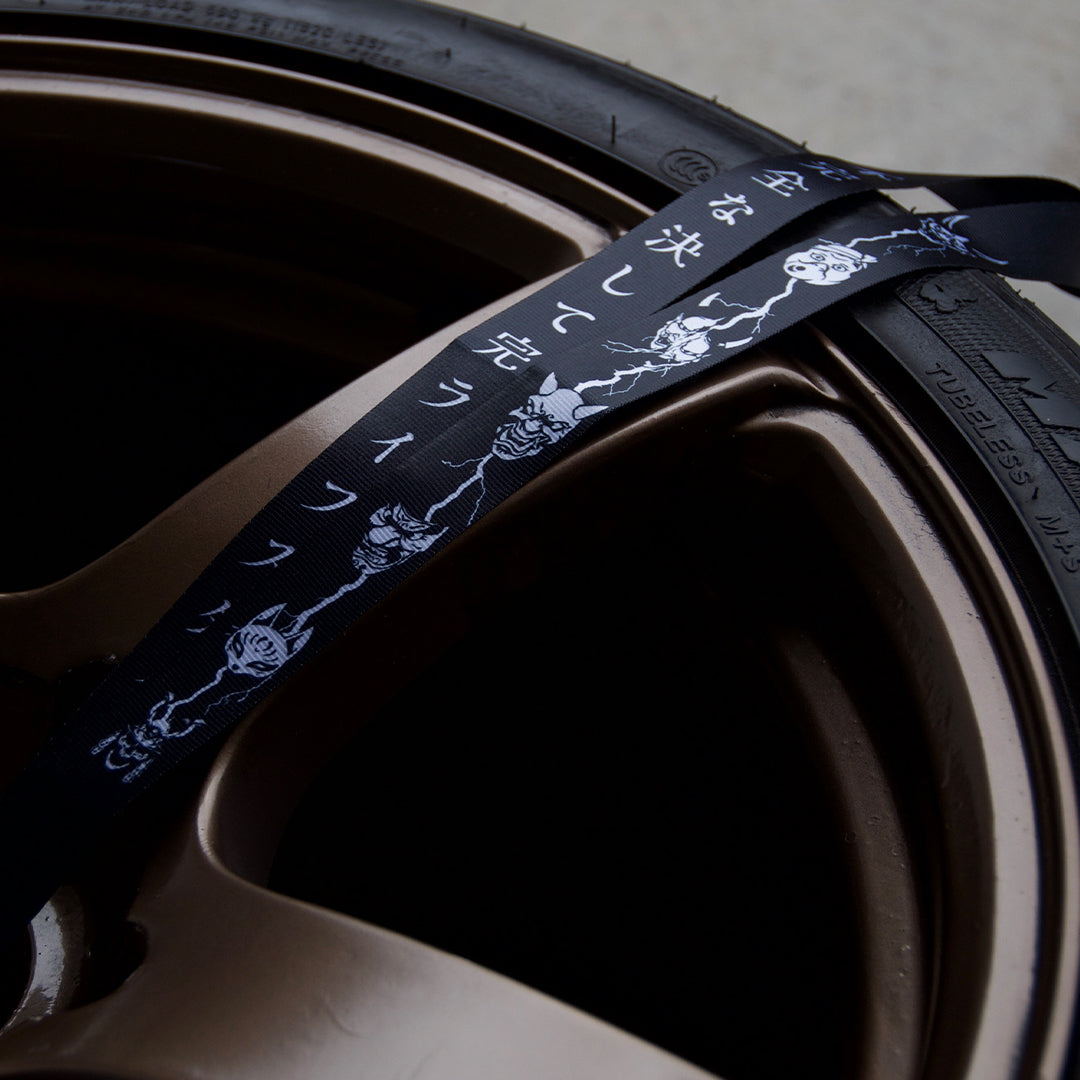 Close-up of a lanyard from The Mask Collection by Incompletegl, crafted from high-quality black nylon featuring white skull designs and Japanese characters, resting on a partially visible bronze-colored car wheel rim. The lighting highlights the metallic texture, perfectly capturing the essence of an automotive lifestyle.