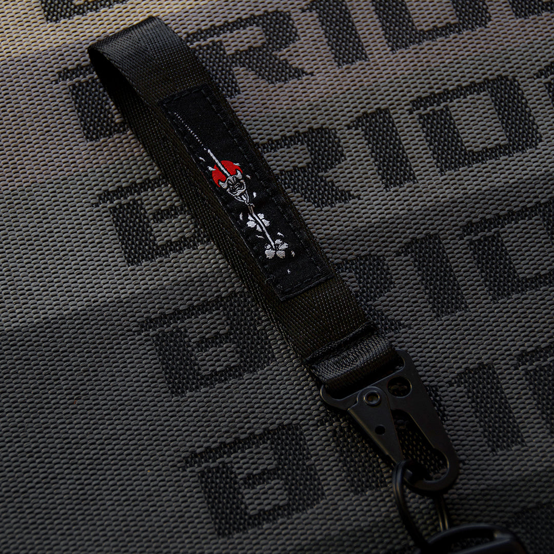 An Oni Mask Sword x Cherry Blossoms carabiner keychain by Incompletegl is displayed on a textured surface featuring a repeating "BRIDE" logo in the background. This black keychain, inspired by Japanese aesthetics, showcases an intricate cartoon skull design with red flame details and includes a carabiner clasp at one end for attaching keys or other items.